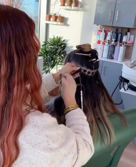 A female stylist with red hair installing hair extensions in her Fort Worth salon suite at Cachet Salons & Spa